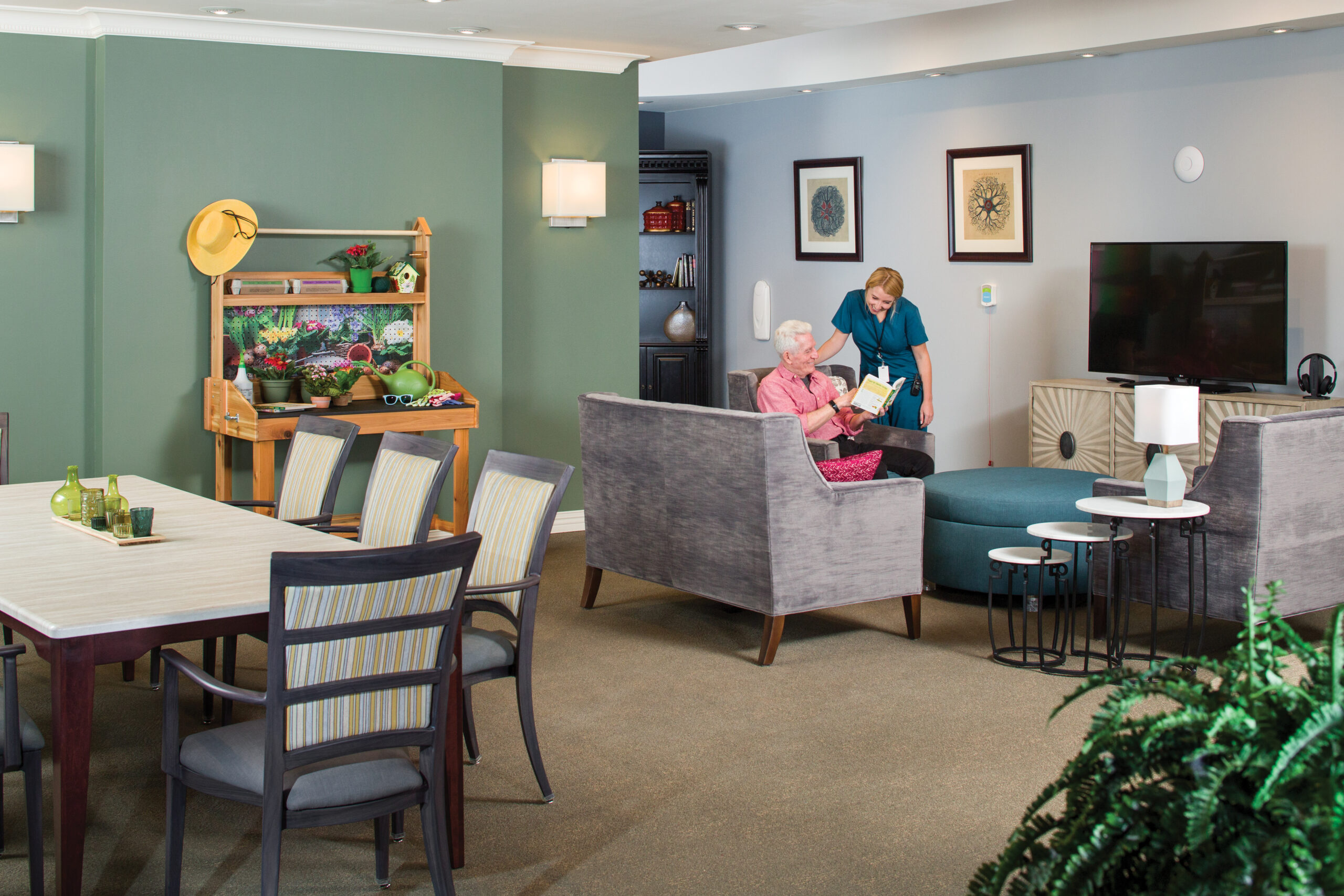 G1376_iu_c Nurse in green scrubs with man in pink shirt in a common room with gardening table