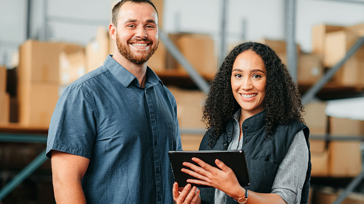 2 people in a warehouse of boxes holding a tablet